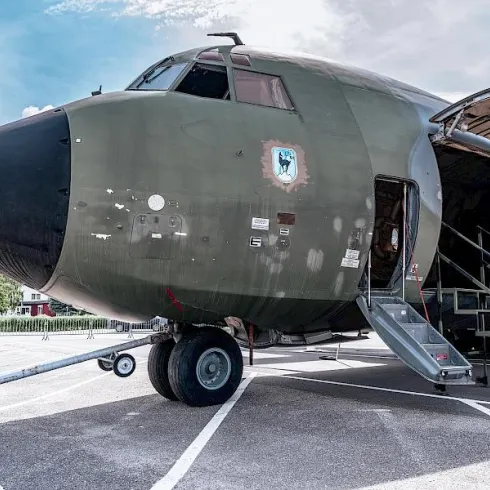 Ein militärisches Flugzeug steht auf einem Parkplatz. Der vordere Teil des Flugzeugs ist geöffnet, und eine Treppe führt hinein.