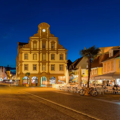 Ein schöner Platz bei Nacht mit historischen Gebäuden und bunten Lichtern. Cafés und Palmen schaffen eine einladende Atmosphäre.