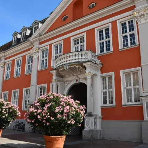 Ein schönes historisches Gebäude mit oranger Fassade und großen Fenstern. Vor dem Eingang stehen gepflegte, blühende Pflanzen in Töpfen.