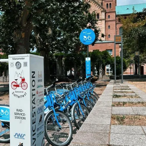 Eine Rad-Service-Station mit blauen Bikes steht auf einem Platz. Im Hintergrund ist ein historisches Gebäude mit einem hohen Turm zu sehen.