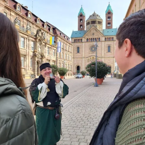 Ein junger Mann in historischer Kleidung spielt ein Instrument vor einem alten Gebäude. Im Vordergrund sind zwei Personen zu sehen, die ihm aufmerksam zuhören.