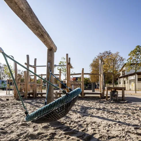 Ein moderner Spielplatz mit einer großen Schaukel und Spielgeräten aus Holz. Der Sandboden und die umgebenden Bäume schaffen eine einladende Atmosphäre.