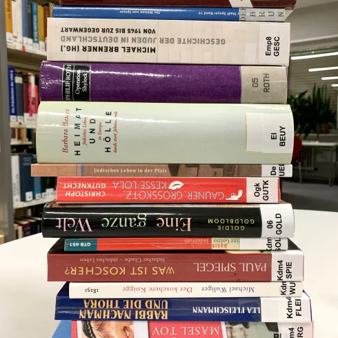 Ein Stapel von bunten Büchern steht auf einem Tisch in einer Bibliothek. Die Buchrücken sind gut sichtbar und zeigen verschiedene Titel und Farben.