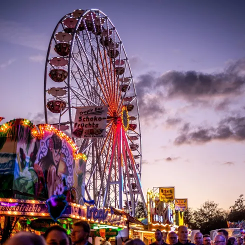 Ein Riesenrad leuchtet am Abendhimmel, umgeben von bunten Fahrgeschäften und festlicher Stimmung. Viele Menschen genießen den Spaß auf dem Jahrmarkt.