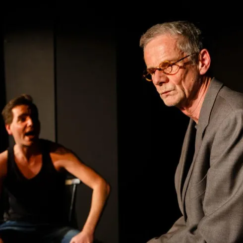 Zwei Männer auf einer Theaterbühne. Der Mann links wirkt lebhaft und emotional, während der Mann rechts ernst und nachdenklich aussieht.