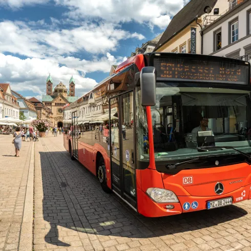 Ein roter Stadtbus steht auf einem belebten Platz. Im Hintergrund sind historische Gebäude und Menschen zu sehen, die die Umgebung genießen.