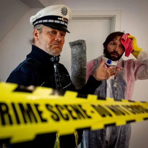 Ein Polizist und ein Mann in Schutzkleidung stehen an einem Tatort. Gelbe Absperrbänder mit der Aufschrift "CRIME SCENE - DO NOT CROSS" sind im Hintergrund sichtbar.