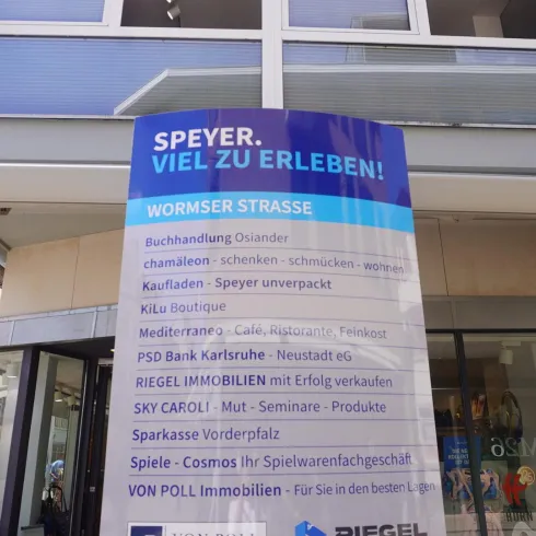 Ein großes Werbeschild in der Stadt Speyer mit verschiedenen Dienstleistungsangeboten. Es zeigt Informationen zu Geschäften und Veranstaltungen in der Wormser Straße.