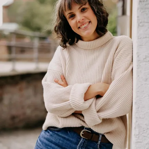 Eine lächelnde Frau steht an einer Wand und trägt einen beigen Pullover und Jeans. Im Hintergrund sind Bäume und eine alte Gebäudestruktur sichtbar.