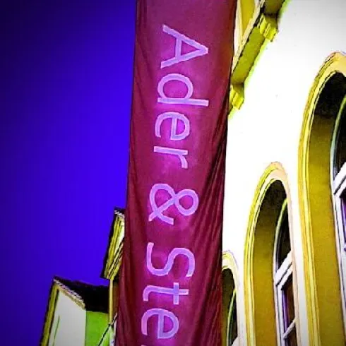 Ein großes Banner mit der Aufschrift „Atelier Ader & Stern“. Es hängt an einem Gebäude in einer bunten Umgebung.
