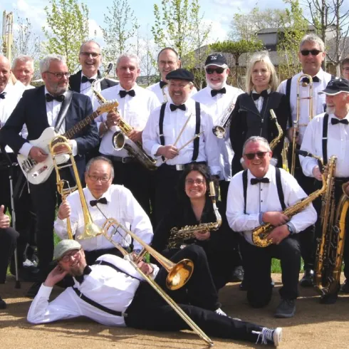 Eine große Gruppe von Musikern steht im Freien und posiert für ein Foto. Sie tragen schwarze und weiße Kleidung und halten verschiedene Instrumente in der Hand.