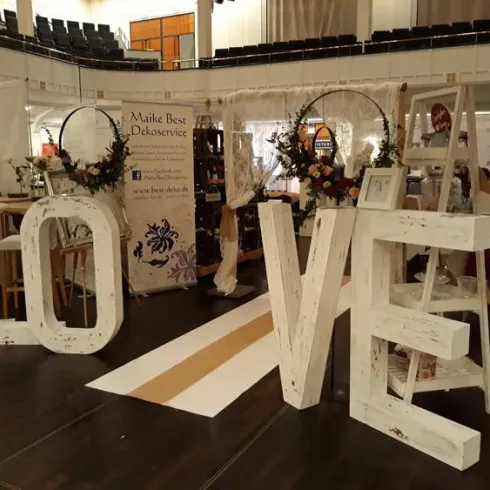 Eine dekorative Art von den Buchstaben "LOVE" aus Holzelementen. Im Hintergrund sind weitere Dekorationen und Stände für eine Veranstaltung zu sehen.