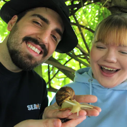 Ein Mann und eine Frau lächeln glücklich, während sie eine große Schnecke halten. Sie befinden sich unter einem grünen Baum.