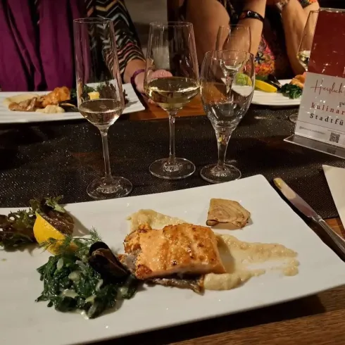 Ein schön angerichteter Teller mit gegrilltem Fisch und Gemüse. Im Hintergrund stehen Gläser mit Wein und man sieht andere Gäste am Tisch.