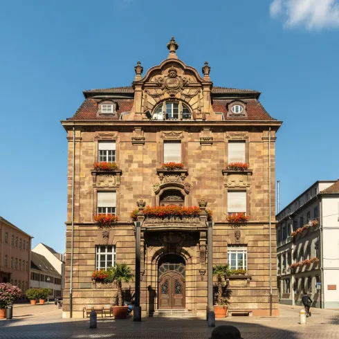 Ein historisches Gebäude mit einer kunstvoll gestalteten Fassade und schönen Blumenkästen. Es steht auf einem Platz, umgeben von weiteren Gebäuden.