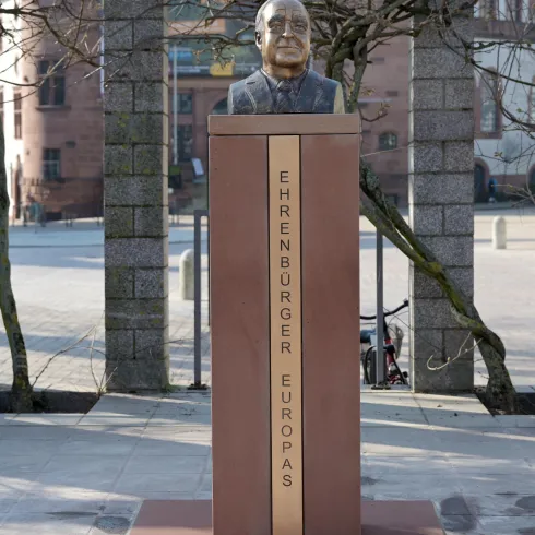 Eine Büste eines Mannes auf einem Sockel mit der Inschrift "Ehrenbürger Europas". Im Hintergrund sind Bäume und Gebäude sichtbar.