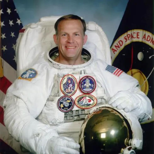 Ein Astronaut in einem Raumanzug lächelt in die Kamera. Im Hintergrund sind Flaggen und Embleme zu sehen.