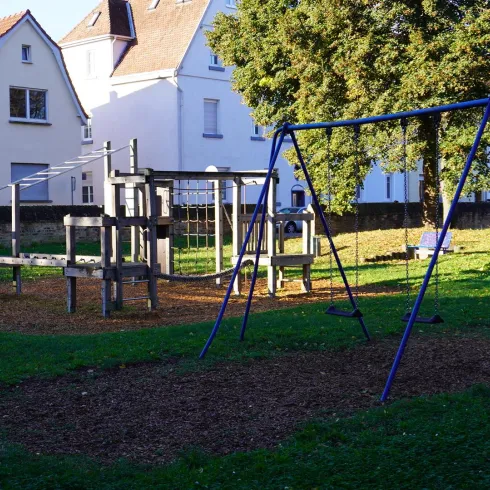 Ein Spielplatz mit einer Rutsche und Schaukeln steht auf einer grünen Wiese. Im Hintergrund sind einige Wohngebäude zu sehen.