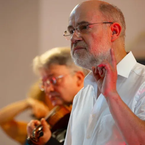 Ein Mann spricht und zeigt mit dem Finger nach oben. Im Hintergrund ist ein Mann mit einer Violine zu sehen.