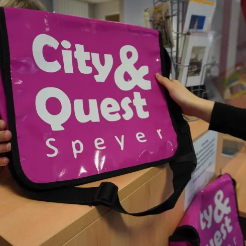 Eine leuchtend pinke Tasche mit dem Aufdruck "City & Quest Speyer" wird an einem Schalter gehalten. Im Hintergrund sind weitere Taschen sichtbar.