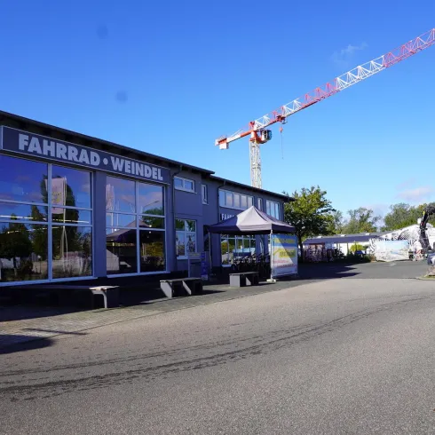 Ein Fahrradgeschäft mit großen Fensterfronten und dem Namen "Fahrrad Weindel" ist zu sehen. Im Hintergrund befindet sich ein Kran und blauer Himmel.
