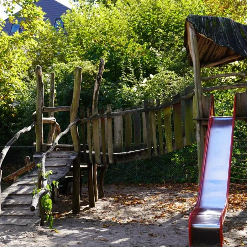 Ein Spielplatz mit einer Holzbrücke und einer roten Rutsche. Umgeben von grünen Bäumen und einem Sandbereich.