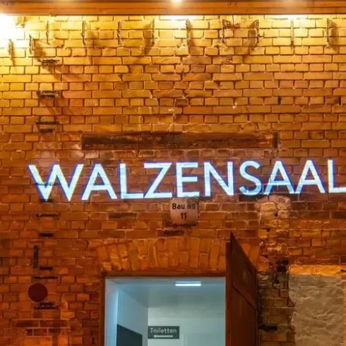 Ein heller Schriftzug „WALZensaAL“ leuchtet an einer Ziegelfassade. Die Eingangstür im Hintergrund ist geöffnet und zeigt einen dunklen Raum.