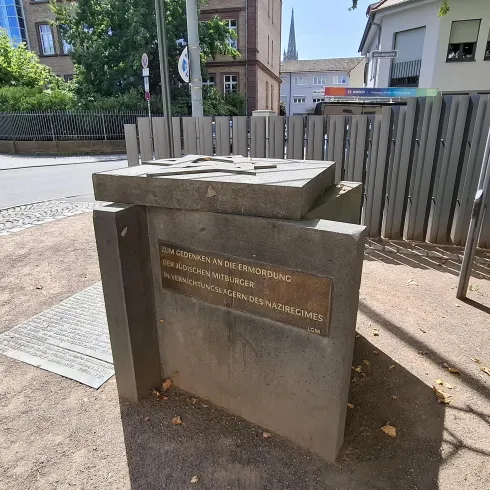 Ein Gedenkstein steht auf einem Gehweg an einer Straße. Die Umgebung ist von Bäumen und modernen Gebäuden umgeben.