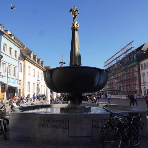 Ein Brunnen im Stadtzentrum umgeben von Geschäften und Cafés. Fahrräder sind an der Seite geparkt, und es herrscht reges Treiben.