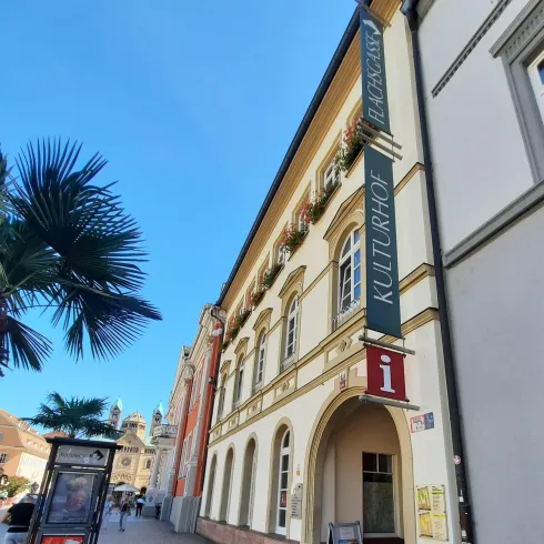 Ein historisches Gebäude mit einer Werbetafel für das Kulturhaus. Im Vordergrund sind einige Palmen und eine entspannte Stadtatmosphäre zu sehen.