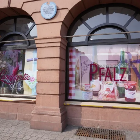 Zwei Schaufenster mit bunten Plakaten und Produkten. Ein Fenster zeigt den Namen "Pfalz" und das andere hebt das Wort "Indulgence" hervor.
