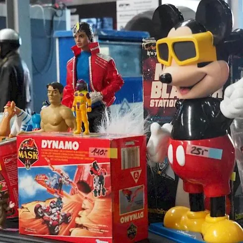 Eine Sammlung von Vintage-Spielzeugen auf einem Tisch. Darunter sind Figuren von Mickey Mouse, He-Man und anderen Charakteren in verschiedenen Farben und Größen.