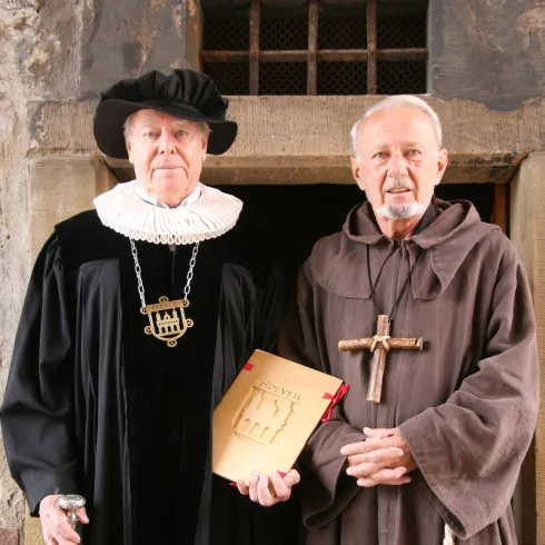 Zwei Männer stehen in historischen Kostümen. Der eine trägt eine schwarze Robe mit einem Kragen, der andere ist in ein braunes Mönchskostüm gekleidet.