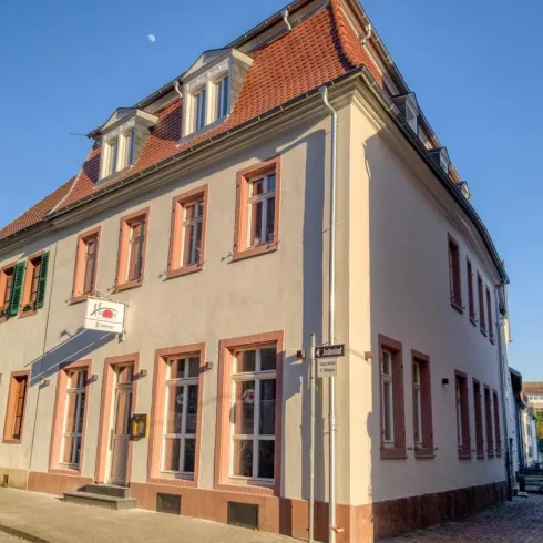 Ein historisches Gebäude an einer Straßenecke mit rot-braunen Fensterrahmen. Das Wetter ist klar und sonnig.