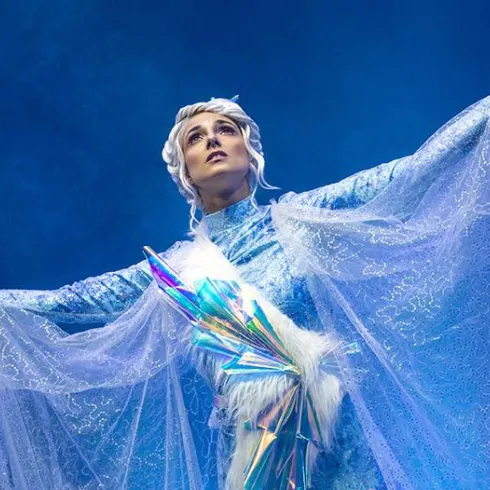 Eine beeindruckende Darstellerin in einem glitzernden, eisblauen Kostüm. Sie breitet ihre Arme aus und strahlt Selbstbewusstsein aus.