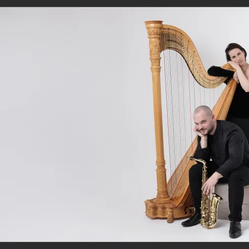 Ein Musiker sitzt mit einem Saxophon auf einem Hocker, während eine Harfenistin hinter ihm steht. Beide tragen elegante schwarze Kleidung und posieren in einem minimalistischen Hintergrund.