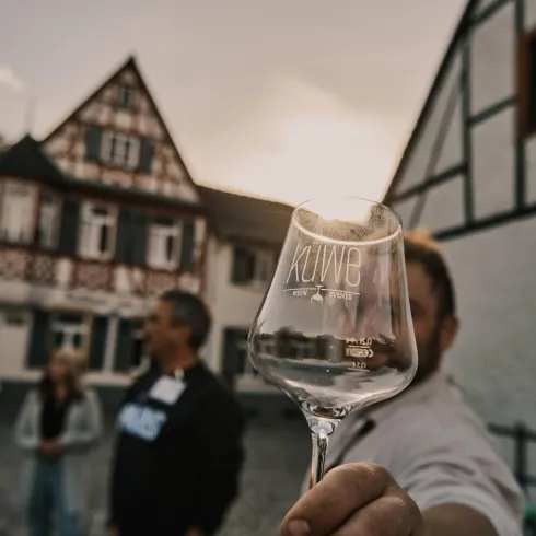Eine Gruppe von Menschen steht vor einem traditionellen Gebäude. Eine Person hält ein Weinglas mit der Aufschrift „Küwe“ in den Vordergrund.