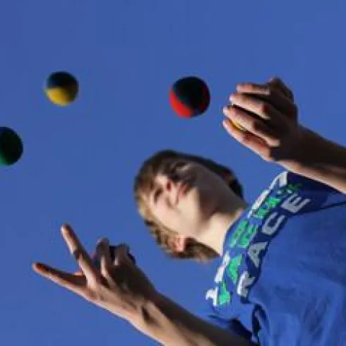 Ein Junge jongliert mit drei bunten Bällen gegen einen klaren blauen Himmel. Er trägt ein blaues T-Shirt und sieht konzentriert aus.