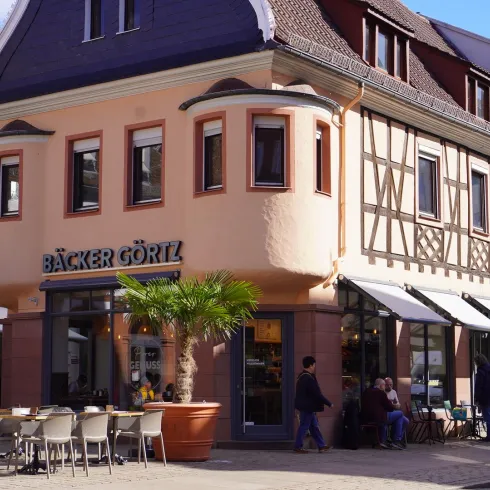Ein charmantes Gebäude mit Fachwerkarchitektur und einem Café namens "Bäcker Görtl". Vor der Fassade stehen Tische und Stühle, umgeben von Palmen.