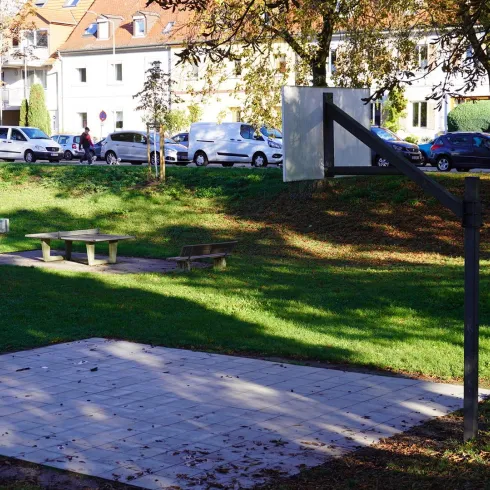 Ein kleiner Park mit einem Basketballkorb und einer offenen Fläche. Im Hintergrund sind parkende Autos und Wohngebäude zu sehen.