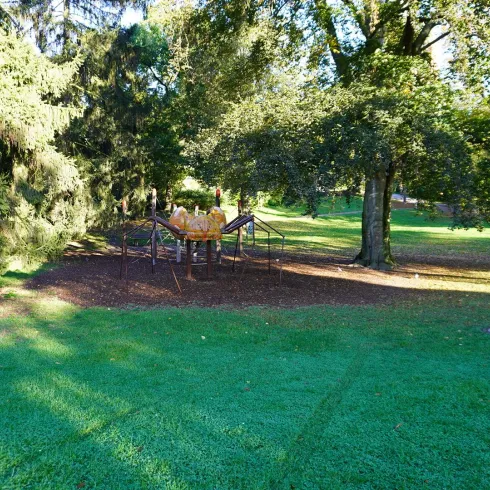 Ein Spielplatz im Park, umgeben von hohen Bäumen. Das Gras ist grün und gut gepflegt.