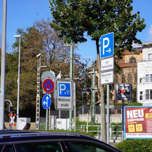 Eine Straßenszene mit vielen Verkehrsschildern, einschließlich Parkmöglichkeiten und einer Bahnübergangsschranke. Im Hintergrund sind Bäume und Wohngebäude sichtbar.