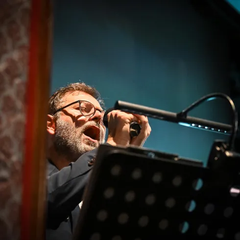 Ein Mann singt leidenschaftlich ins Mikrofon. Er steht vor einem Notenständer mit beleuchtetem Text.