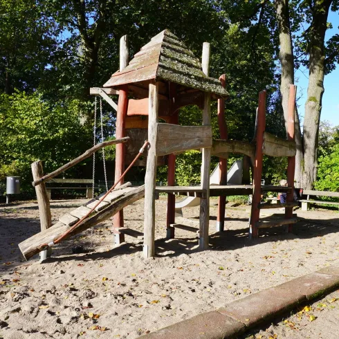 Ein Spielplatz mit einem hölzernen Spielgerüst und einer Rutsche. Der Bereich ist von Bäumen umgeben und hat Sandboden.