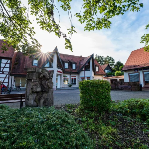 Ein malerischer Platz mit traditionellen Fachwerkhäusern und einer schönen grünen Umgebung. Die Sonne scheint durch die Bäume und sorgt für eine angenehme Atmosphäre.