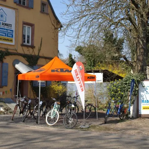 Ein Fahrradgeschäft mit einem orangefarbenen Zelt und verschiedenen Fahrrädern steht vor einem Gebäude. Neben dem Eingang gibt es farbige Fahnen und Werbung für das Geschäft.
