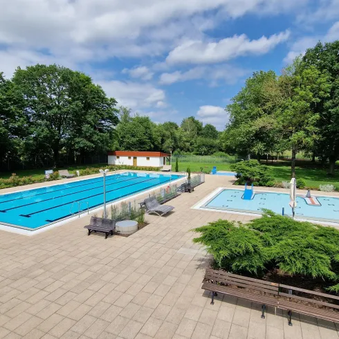 Ein Freibad mit einem großen Sportbecken und einem Kinderbecken. Umgeben von Bäumen und einer grünen Rasenfläche.