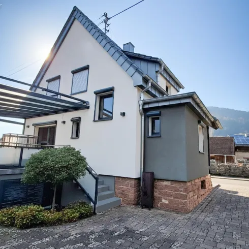 Ein modernes Haus mit einer Terrasse und einer klaren Fassade. Im Hintergrund sind sanfte Hügel und ein blauer Himmel zu sehen.