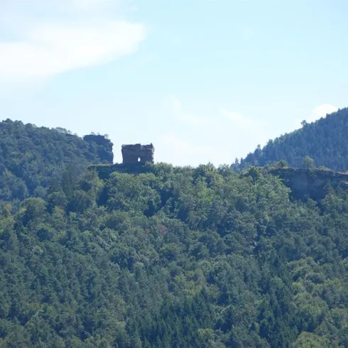 Eine alte Ruine auf einem Hügel, umgeben von grünen Bäumen. Der Himmel ist blau und klar.