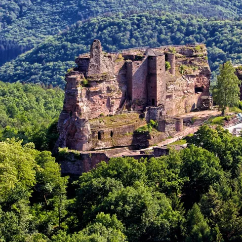 Eine beeindruckende Burgruine steht auf einem grünen Hügel, umgeben von dichten Wäldern. Im Hintergrund erstreckt sich eine malerische Landschaft mit sanften Hügeln.